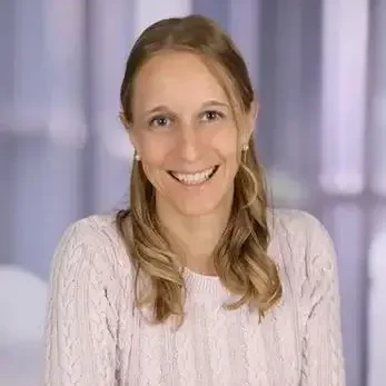 Lächelnde Frau mit langen, blonden Haaren in einem hellen Pullover vor verschwommenem Hintergrund.