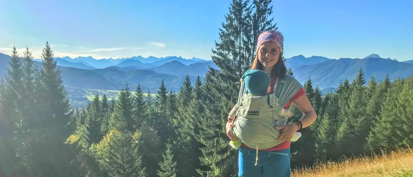 Frau mit Baby-Trage vor einer bergigen Landschaft.