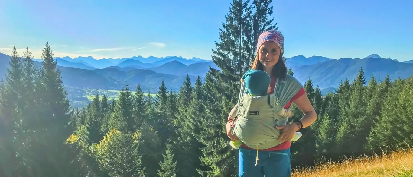Frau mit Baby-Trage vor einer bergigen Landschaft.