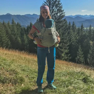 Angelique Rest mit Baby im Tragetuch, auf grüner Wiese vor einer Bergkulisse.