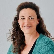 Frau mit lockigem Haar, freundlichem Lächeln und grüner Jacke vor neutralem Hintergrund.