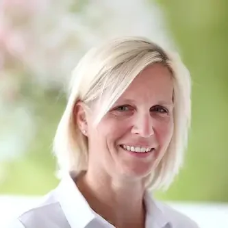 Frau mit blonden Haaren und freundlichem Lächeln vor unscharfem, hellem Hintergrund.