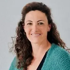 Frau mit lockigem, braunem Haar und grünem Pullover, lächelt freundlich.